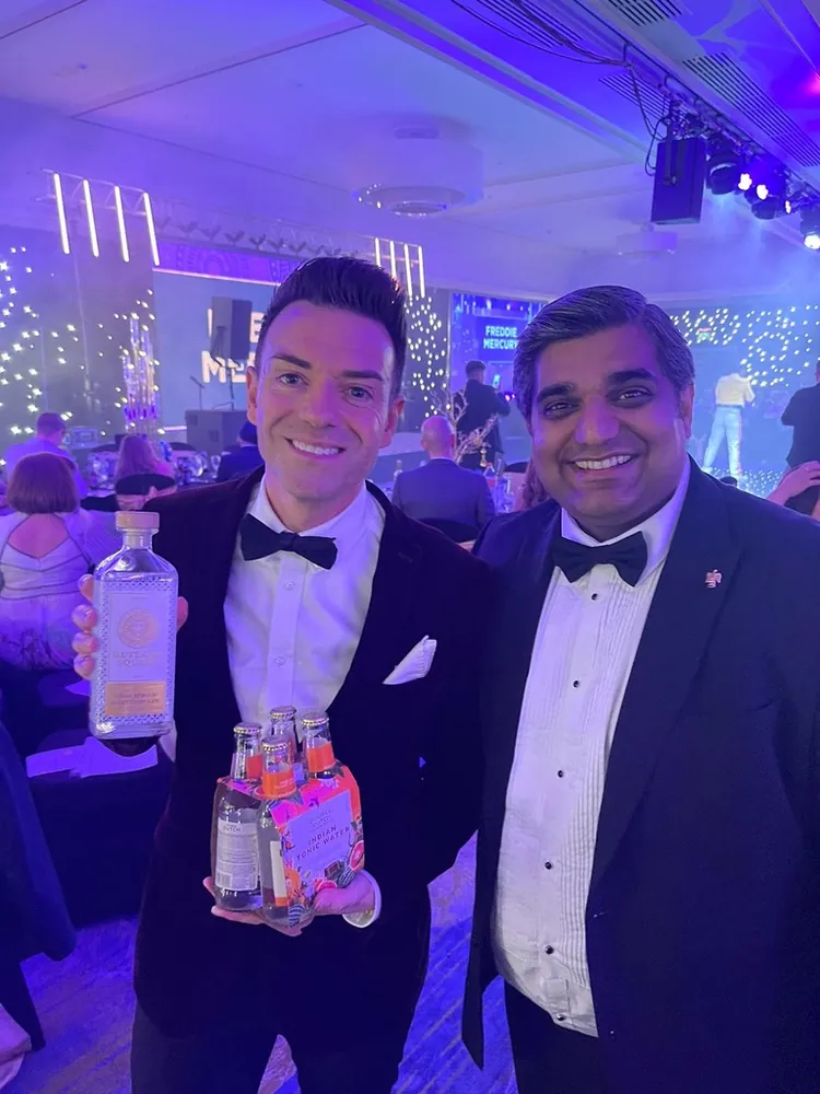 A man in a black dinner jacket and bow tie holds a bottle of Rutland Square gin and a pack of tonic beside another man in black tie, under blue and purple lighting