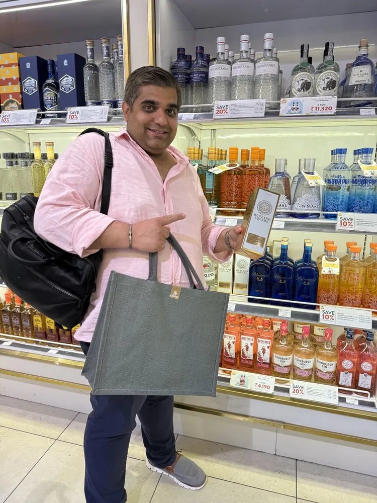 A man in a pink shirt points to a Rutland Square gift box among bottles on a duty-free shelf, holding a grey bag