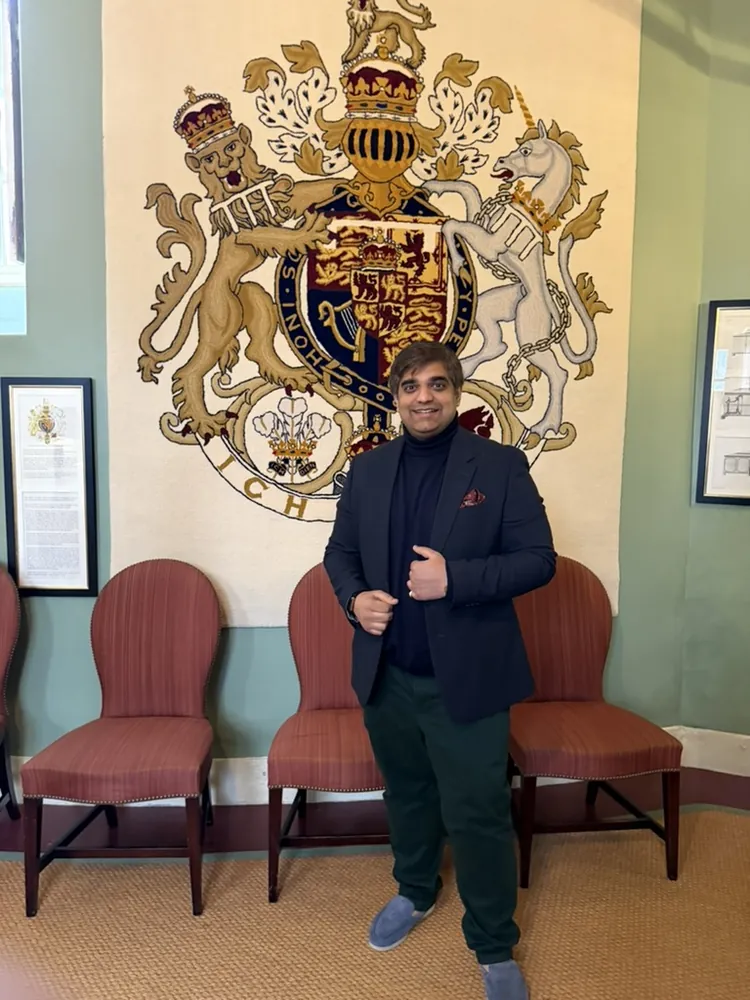 A man in a navy jacket stands in front of a large embroidered coat of arms hung on a wall, beside red chairs