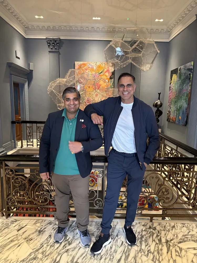 Two men standing together on the landing of an ornate staircase, in a room hung with paintings