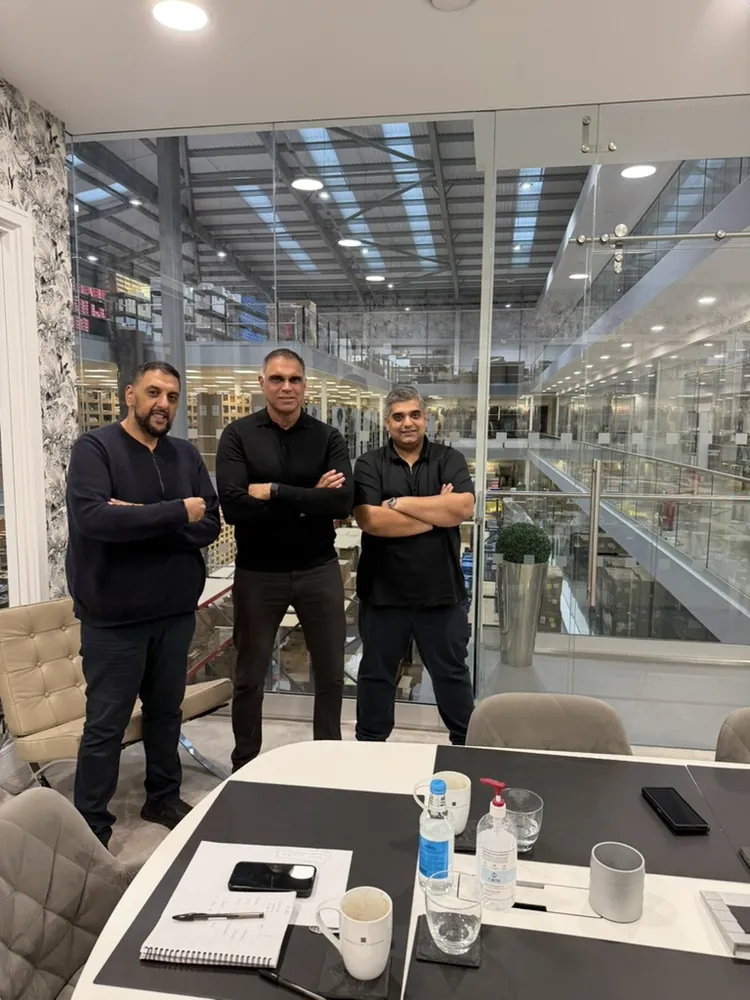 Three men standing with folded arms beside a meeting table, in front of a glass wall that overlooks a large indoor space