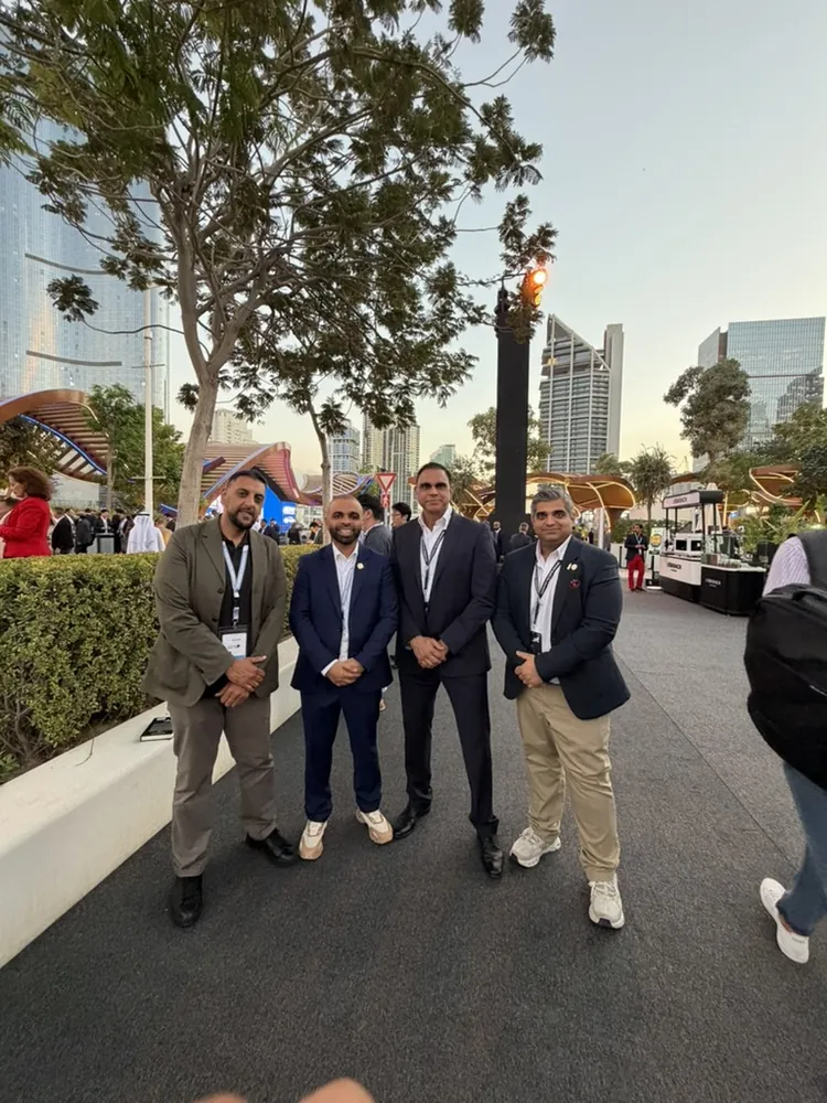 Four men standing together on a paved path outdoors, with trees and tall buildings behind them