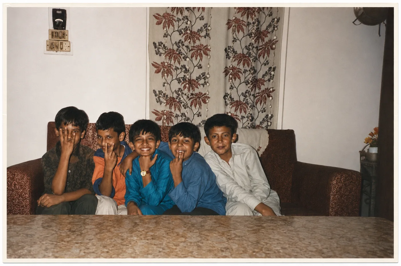 Five boys on a sofa in front of floral wallpaper, in a colour-graded family photograph with a white print border: one hides his face behind his hand, two make V-signs beside their mouths and the others smile; Nishant Sharma is the middle boy in the blue shirt, with his hand on his chin