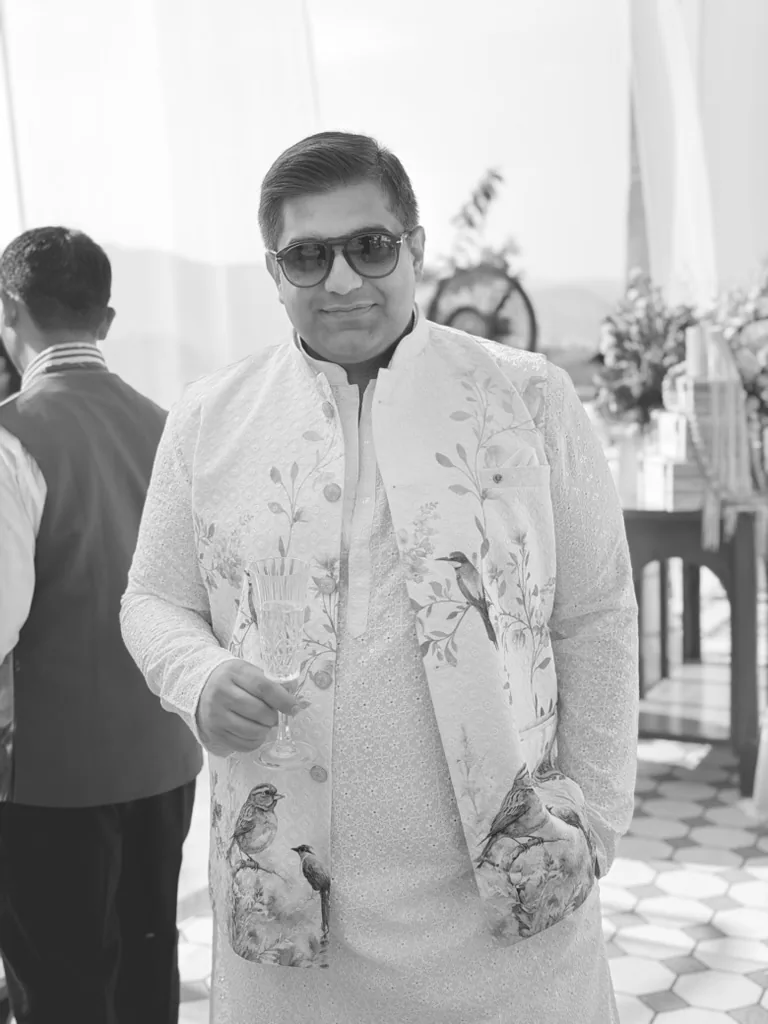 A man in black and white, in sunglasses and a white embroidered kurta with a bird-print waistcoat, holding a glass