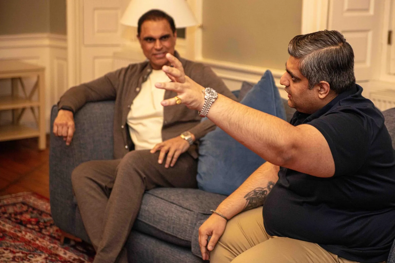 Nishant Sharma gesturing as he talks with Nick Bassi, who sits back on a grey sofa in a panelled room