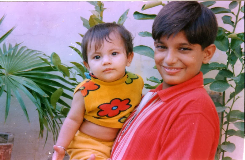 A smiling boy in a red shirt holding a baby in a yellow flowered top in front of palm and lemon leaves