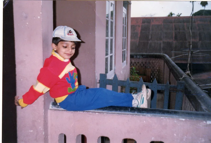 A small boy in a white cap, a red and yellow jumper and blue trousers sitting on a pink balcony wall, smiling down at his feet, with a corrugated roof and palm trees behind