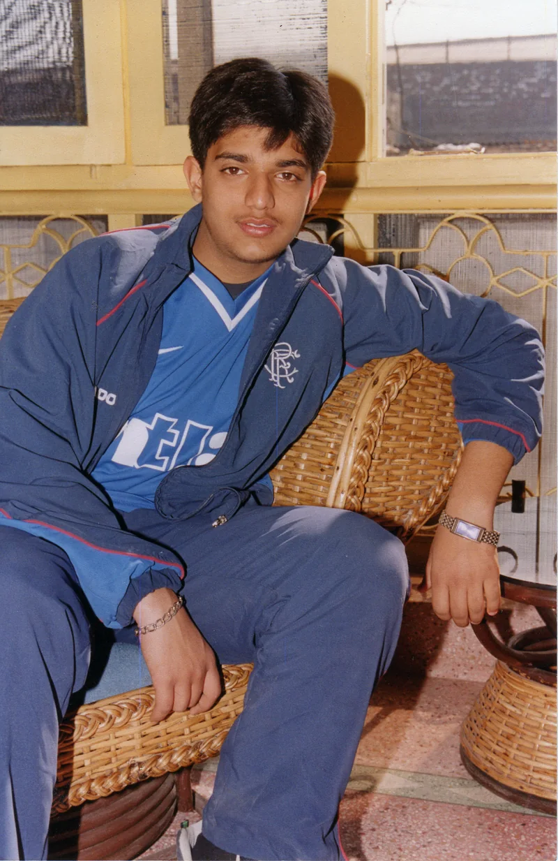 A teenager in a Rangers football shirt and navy tracksuit sitting in a wicker chair by yellow window frames, wearing a wristwatch and a bracelet