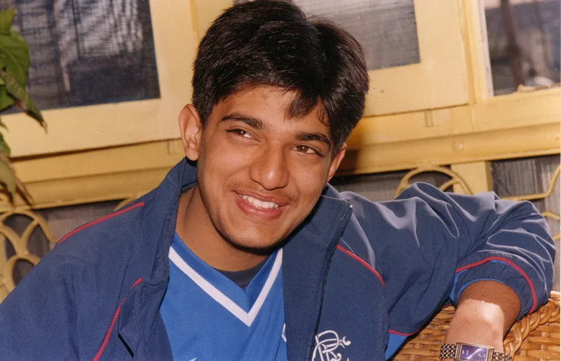 A close-up of a laughing teenager in a navy Rangers tracksuit jacket over a blue football shirt, leaning on a wicker chair
