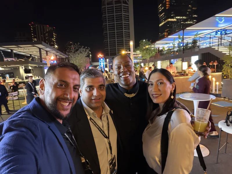 Four people with event lanyards taking a smiling selfie at a night-time terrace event, with lit towers behind