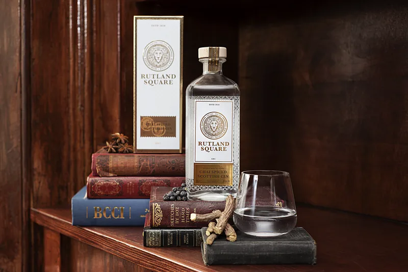 A bottle of Rutland Square Chai Spiced Scottish Gin and its white and gold carton on a stack of old leather-bound books, with star anise, juniper berries, liquorice root and a tumbler of gin, against dark wood panelling