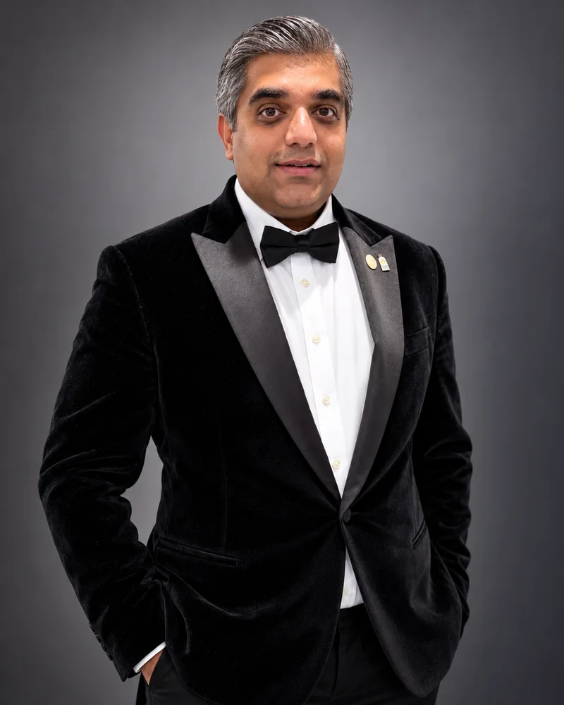 Nishant Sharma in a black velvet dinner jacket with satin peak lapels, a white shirt and black bow tie, two pins on the lapel, against a grey studio background