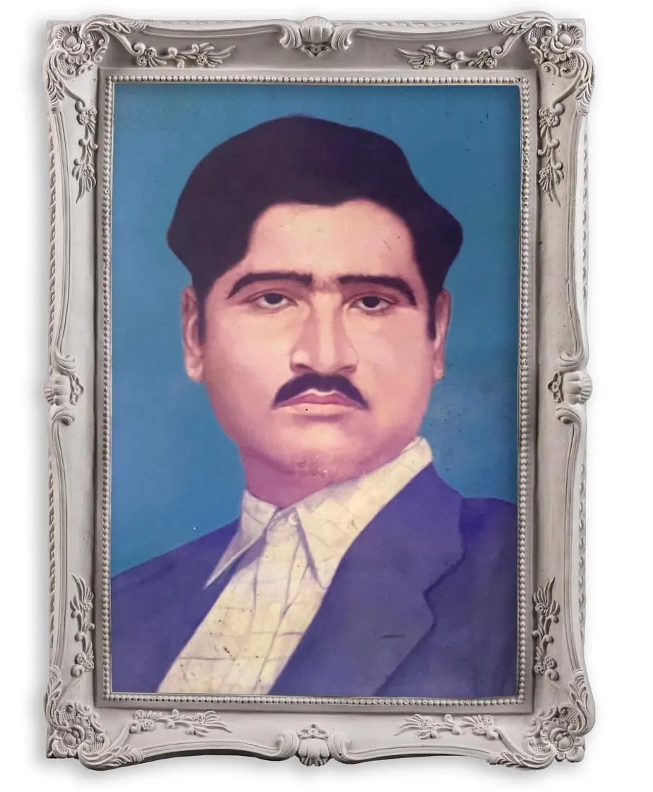 Hand-tinted portrait of Tej Ram Bawa in an ornate silver-grey frame: a man with dark hair and a moustache, in a blue jacket and a light patterned shirt