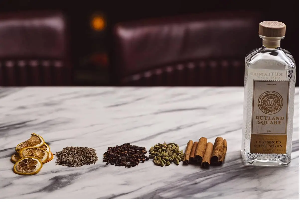 A bottle of Rutland Square gin beside a row of botanicals laid on marble: dried orange slices, seeds, spices and cinnamon sticks