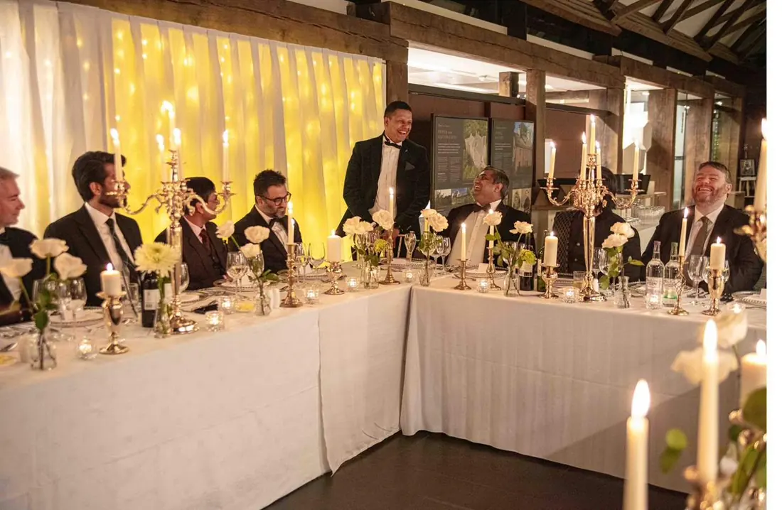 Guests seated along a long table dressed in white linen, with candelabra and candles, in a hall with a timber roof and a curtain hung with fairy lights