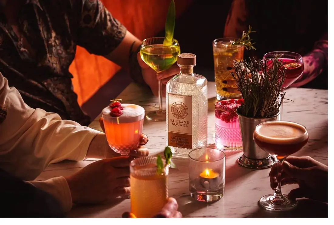 Cocktails and a bottle of Rutland Square gin on a candlelit marble table, with hands reaching in