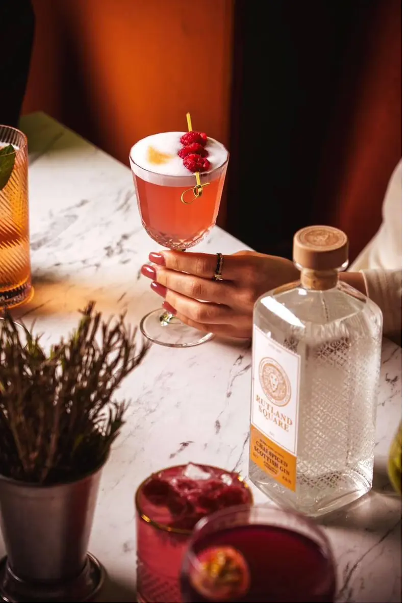 A hand holding a coupe of red cocktail beside a bottle of Rutland Square gin on a marble table, with a sprig of heather in a metal cup