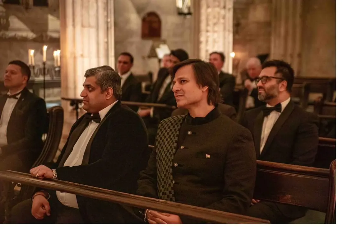Men in dinner jackets seated on benches in a stone hall with arched windows and lit candles