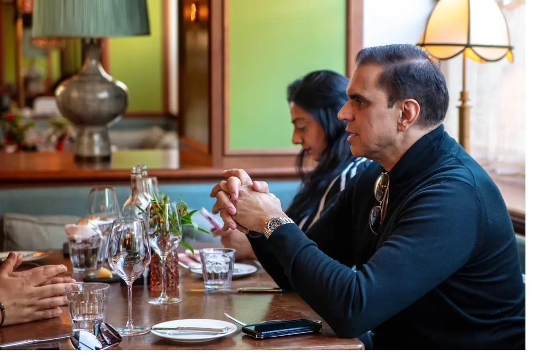 A man in a dark sweater with his hands clasped, talking at a restaurant table, with a woman seated behind him