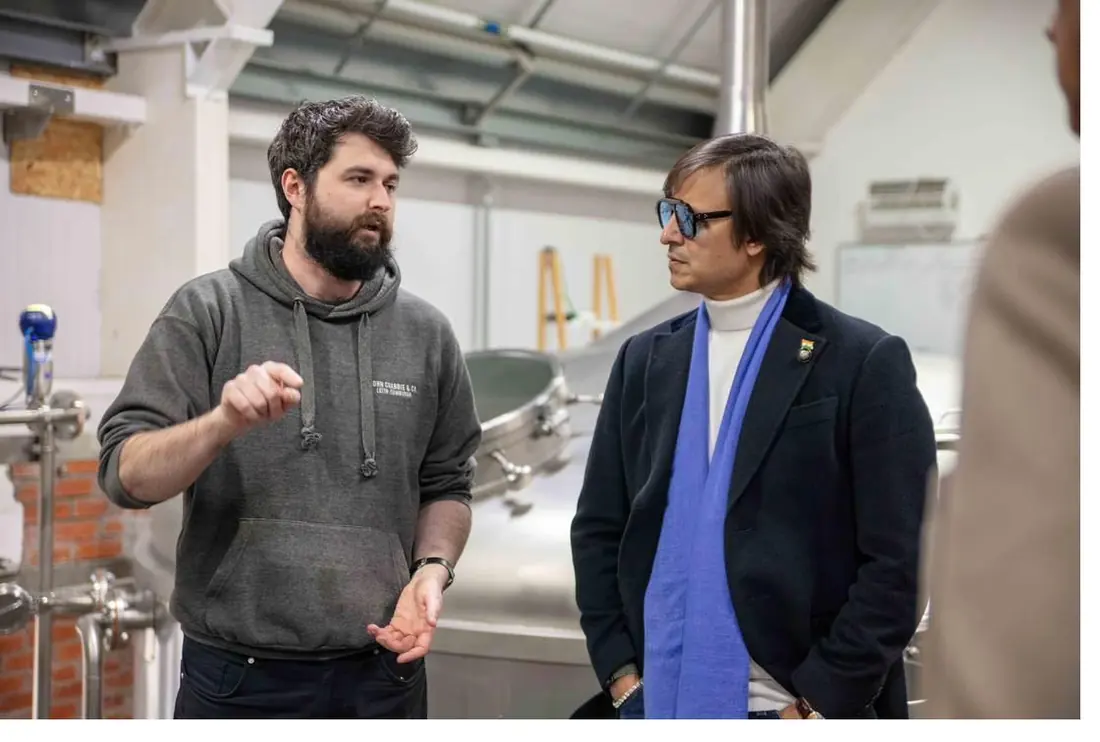 Two men in conversation beside stainless steel equipment: one bearded, in a grey hooded top, the other in a dark jacket with a blue scarf