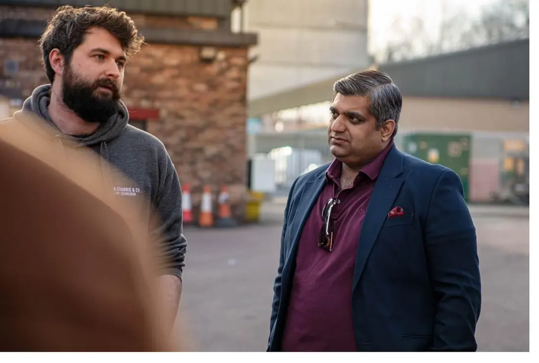 Two men talking in a yard beside a brick building: one bearded, in a grey hooded top, the other in a dark jacket and a maroon shirt