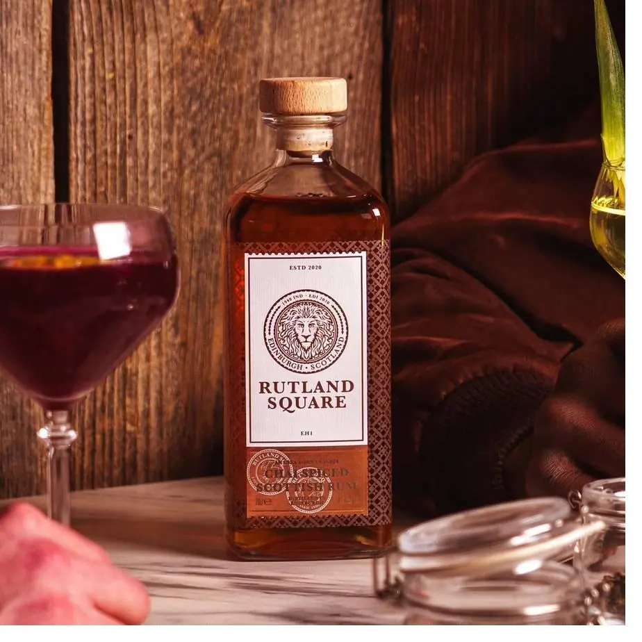 A bottle of Rutland Square Chai Spiced Scottish Rum on a wooden table beside a glass of red drink