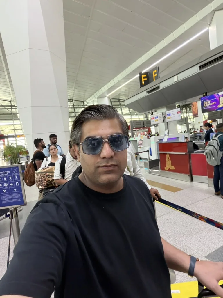 A selfie in blue-tinted sunglasses and a black T-shirt in a bright airport check-in hall, with travellers queuing behind