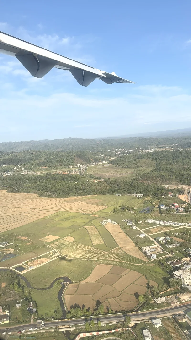 An aircraft wing above green hills, patchwork fields, a winding stream and scattered houses, seen from a plane window