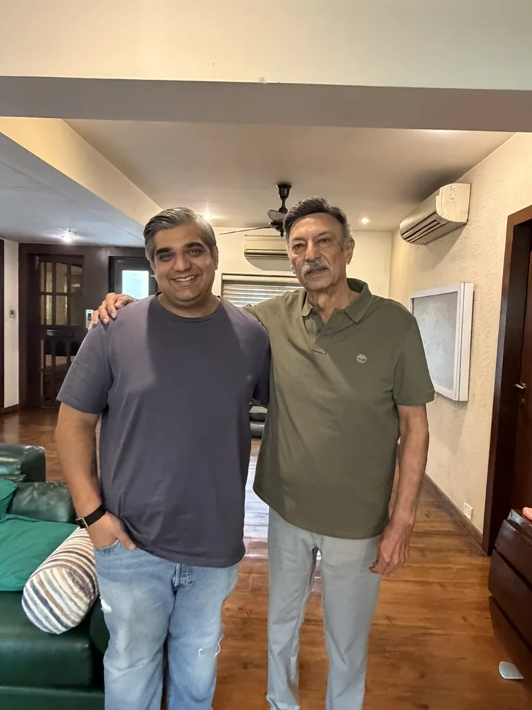 Two men smiling with arms around each other in a living room with wooden floors and a green leather sofa