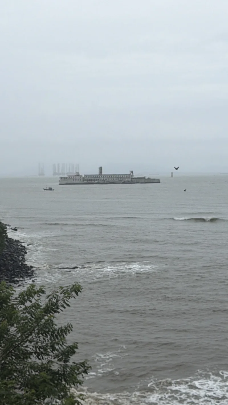 A grey sea in monsoon mist with a long stone structure offshore, a small boat and distant rigs, framed by rocks and leaves