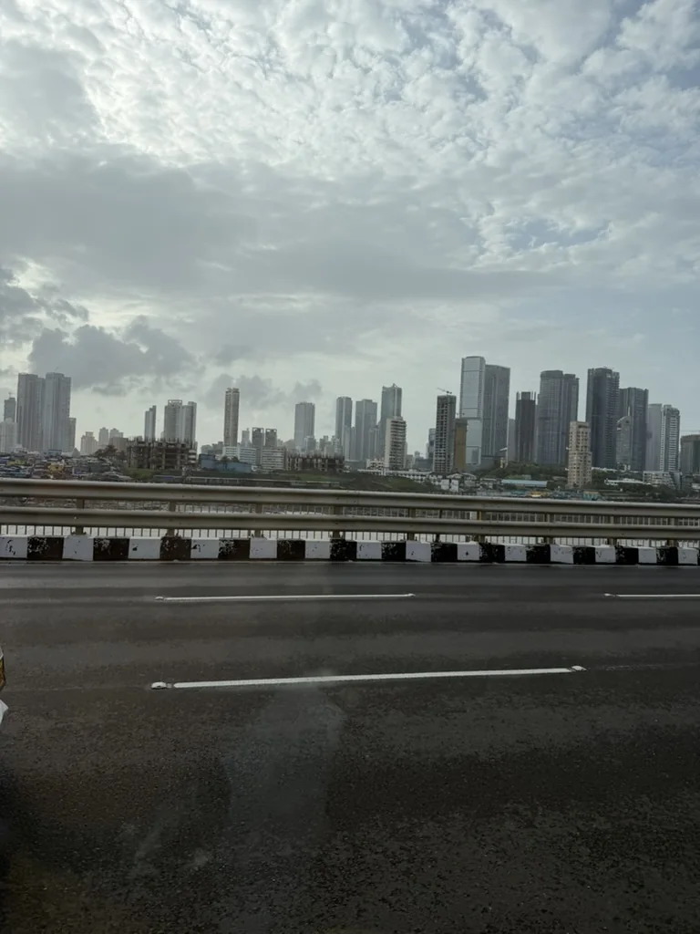 A city skyline of tall towers under a clouded sky, seen across water from a road bridge