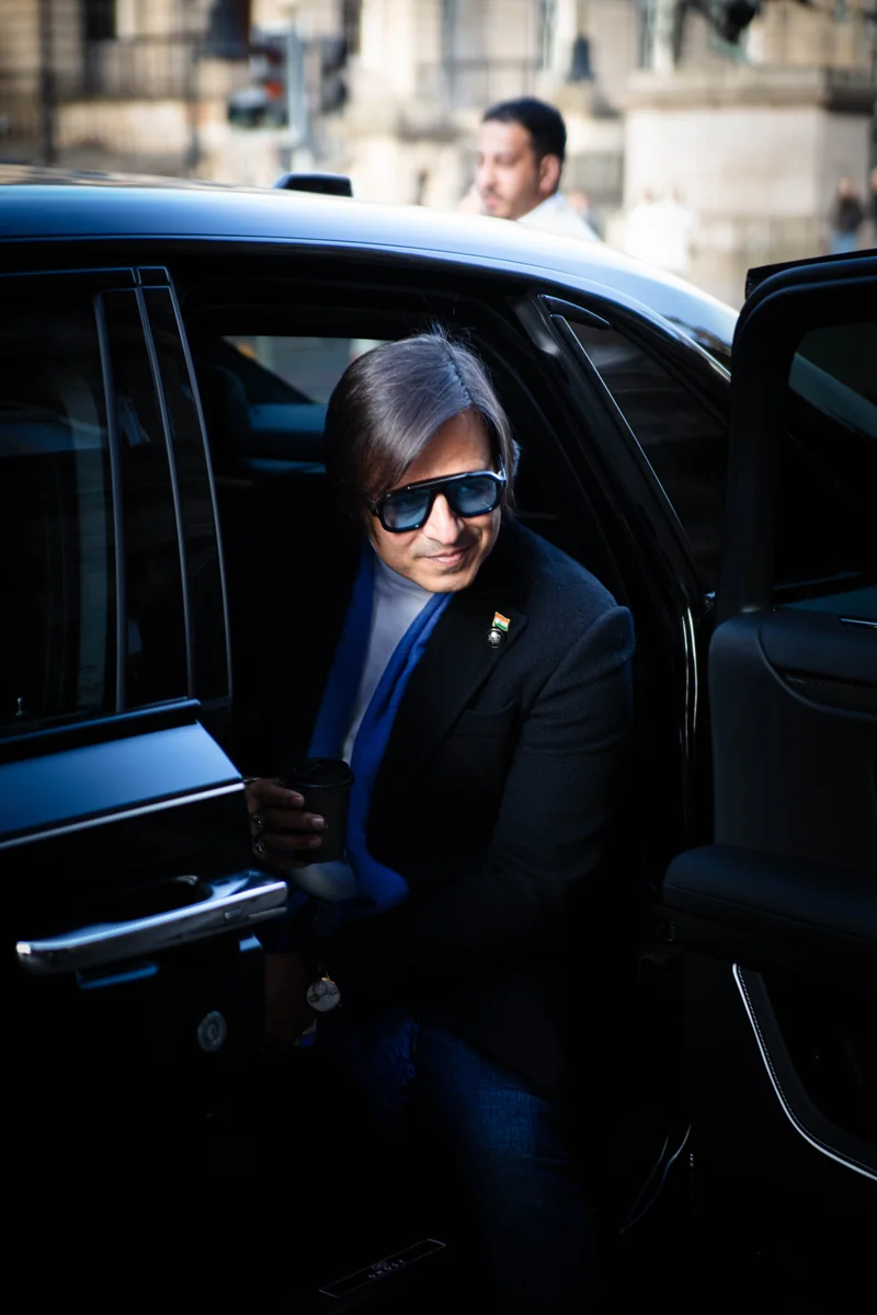 A man in blue-tinted sunglasses, a dark coat and a blue scarf stepping out of a black car with a coffee cup in hand