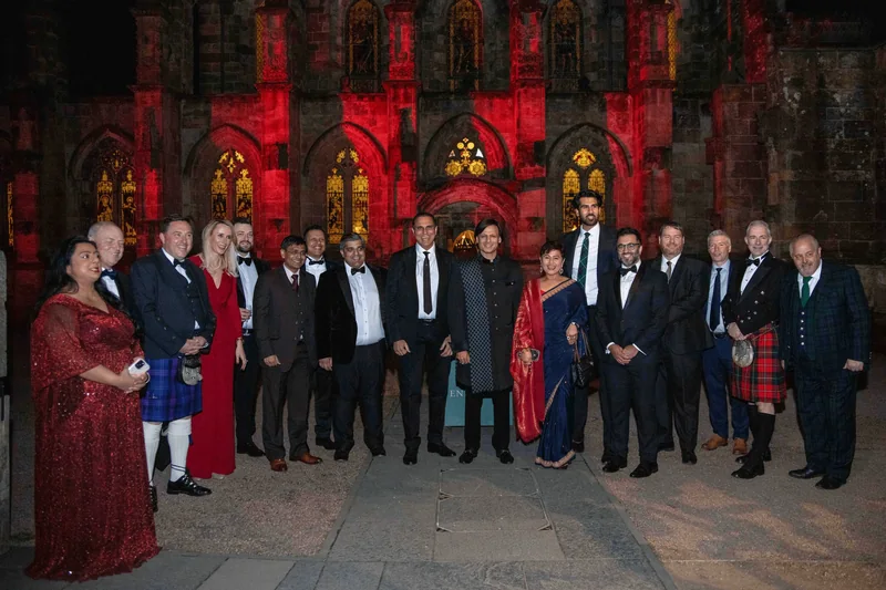 About twenty guests in black tie, kilts, saris and evening dresses standing together outside a Gothic chapel floodlit in red, with lit stained-glass windows