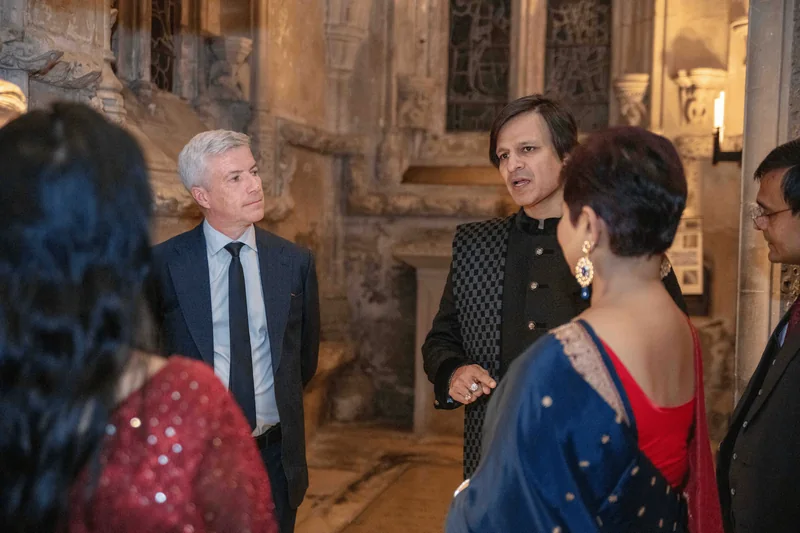 A man in an embroidered black jacket talking with guests among carved stone walls and stained glass
