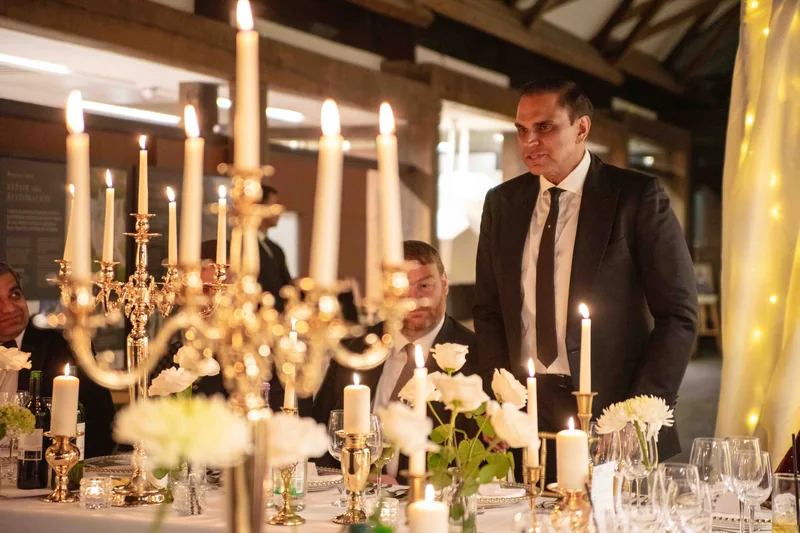 A man in a dark suit standing to speak at a long dinner table set with tall candelabra, white roses and glassware