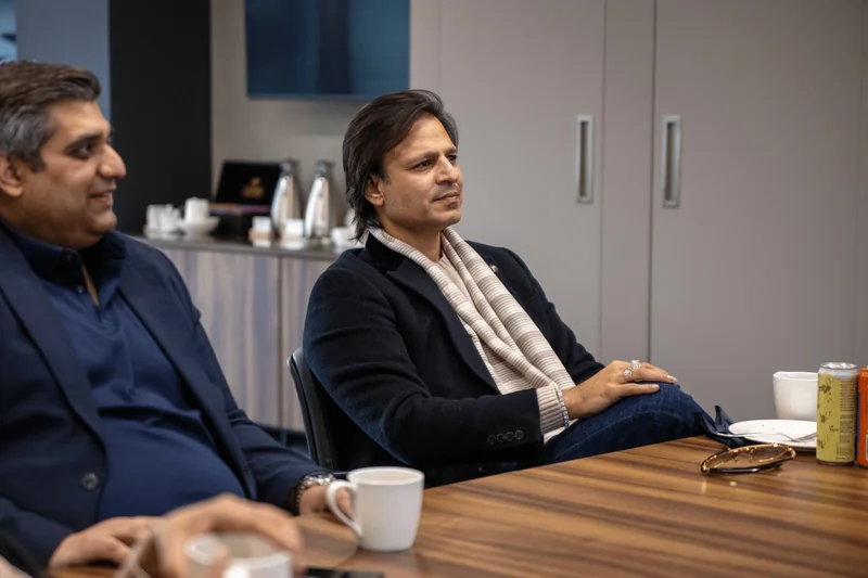 Two men seated at a wooden meeting table with coffee cups, one in a navy jacket and a cream striped scarf