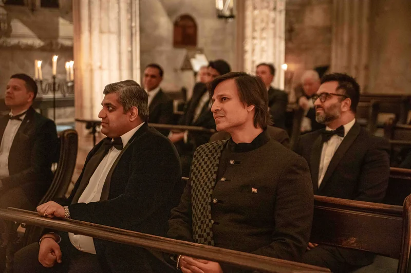 Guests in black tie and an embroidered jacket seated in wooden pews in a stone chapel lit by candles