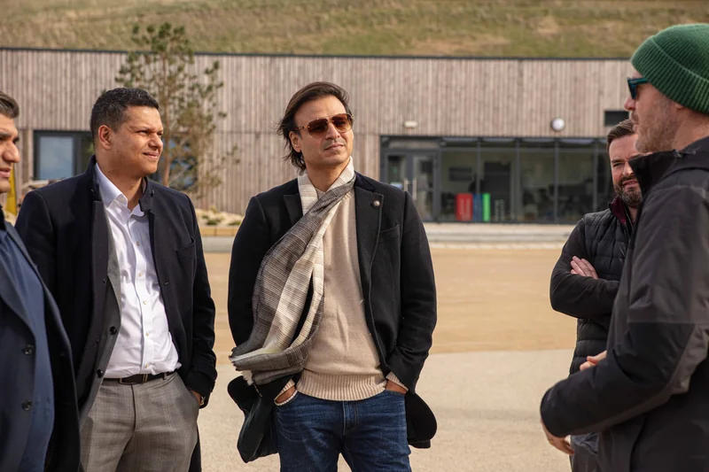 A group in coats talking outside a long timber-clad building beneath a grassy bank, one man in sunglasses and a striped scarf
