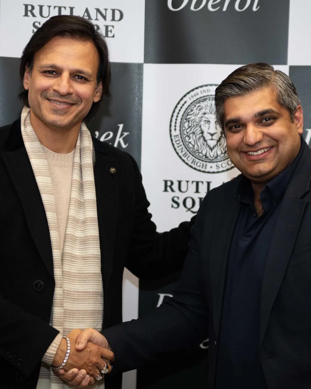 Dr Vivek Anand Oberoi, in a black coat and striped scarf, shakes hands with Nishant Sharma in front of a Rutland Square backdrop
